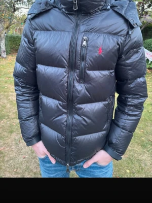 Svart pufferjacka med huva från Ralph Lauren - Säljer en svart pufferjacka med huva och dragkedja framtill. Jackan har en bröstficka med dragkedja och en liten röd logga på bröstet. Fylld med vaddering för extra värme och har breda, quiltade paneler. Perfekt för kalla vinterdagar. Nypris 7000kr, mitt pris 2300kr