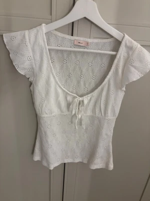 Vit blus med spets och knyt från Nelly - Söt vit blus från Nelly med broderade spetsdetaljer och korta, fladdriga ärmar. Toppen har en djup v-ringning med knyt framtill och är figurnära i modellen. Perfekt för sommarens alla tillfällen!