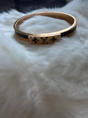 Guldigt armband  - Snyggt armband i guldfärgad metall med svart detalj och logga framtill. Stelt och elegant med en exklusiv känsla, perfekt för dig som gillar lyxiga accessoarer och vill sticka ut lite extra.