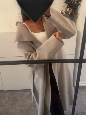 Beige kappa med skärp och bred krage - Snygg beige kappa med bred krage och knytskärp i midjan. Kappan har en rak passform och är tillverkad i ett mjukt, ull-liknande material. Perfekt för kyligare dagar och ger en stilren look.