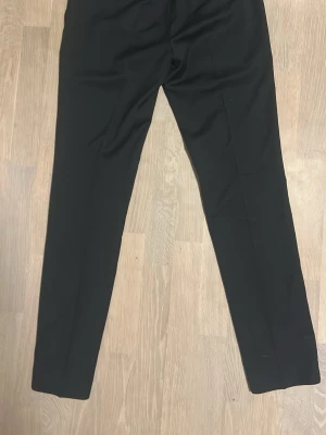 Svarta klassiska kostymbyxor - Säljer ett par svarta kostymbyxor med rak passform och pressveck. Byxorna har en stilren look och passar perfekt till en dressad outfit. Materialet känns mjukt och har en lätt stretch för extra komfort.