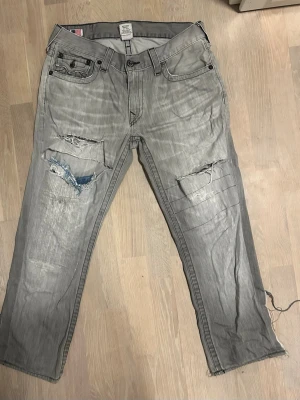 Gråa true religion jeans - Gråa slitna true religion jeans, klippte dem bootcut för att jag ville ha mer swag. midjan är stor så föredrar bälte med. de har hål o har blivit sydda men ändå feta. De är äkta men yeah de ser ju helt fucked ut ahahaha. kan sänka till 50 och kan gärna byta till något annat sjyst. behöver pengar till snus o kläder!