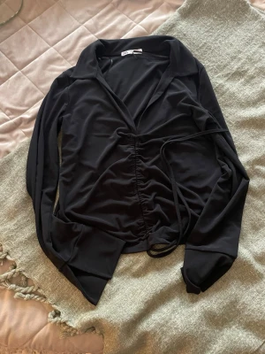 Svart omlottblus från Zara - Snygg svart omlottblus från Zara med långa ärmar och krage. Blusen har knytband i midjan och lätt rynkad effekt framtill. Tillverkad i ett mjukt, stretchigt material som ger en skön passform och trendig look.