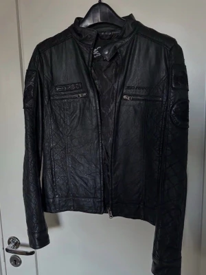 ED Hardy jacka Svart skinnjacka storlek s -  ooos detta är på ett ungerfär sen kan ed hardys kläder skilja sig från våran storlekar även om det står s för det har jag märkt på andra ed hardy kläder som vi haft. Storlek S 34/36  Svart jacka med dragkedja framtill och ståkrage. Jackan har två fickor med dragkedja framtill i brösthöjd. Ärmsluten har dragkedjor för justering. Baksidan av jackan pryds av ett stort, detaljerat motiv med text och en dödskalle. Mindre detaljerade motiv finns även på ärmarna. Insidan av jackan har ett mönstrat 