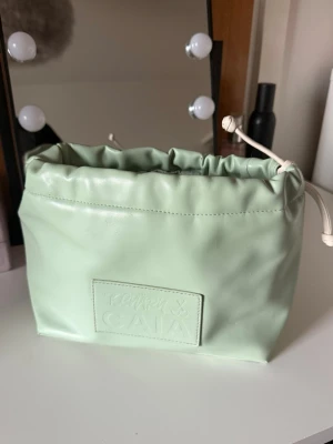 Mintgrön necessär från CAIA x Tchary - Snygg mintgrön necessär från CAIA x Tchary i mjukt konstläder. Väskan har dragskostängning upptill och en stilren patch med logga på framsidan. Perfekt för smink eller småprylar när du är på språng.