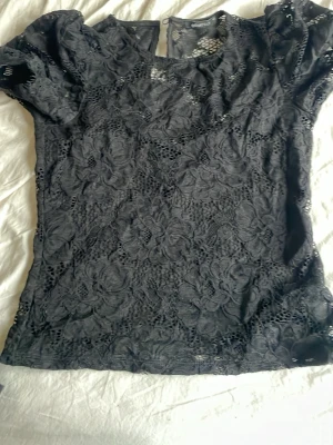 Svart spetsblus med blommönster - Säljer en svart blus i spets med blommigt mönster. Toppen har korta ärmar och är lätt transparent med fina detaljer. Perfekt att styla med jeans eller kjol för en trendig look.