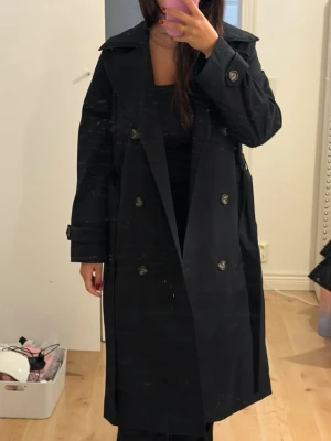 Svart trenchcoat med knytskärp - Stilren svart trenchcoat med dubbelknäppning och brett slag. Knytskärp i midjan och dekorativa knappar vid ärmslut. Lång modell som ger en klassisk och trendig look. Perfekt för lager-på-lager och funkar till många outfits.