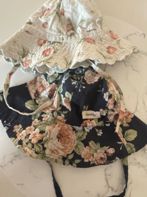 Blommiga solhattar från Newbie - Två söta solhattar från Newbie med blommigt mönster. En hatt är ljus med rosa blommor och vågig kant, den andra är mörkblå med stora blommor i beige och grönt. Båda har knytband under hakan och är perfekta för soliga dagar.