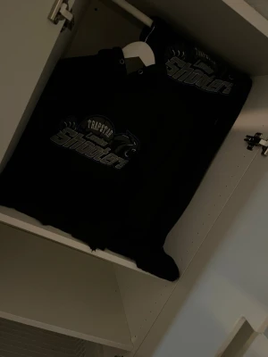 Svart hoodie Trapstar Shooter - Svart hoodie med trycket 'Trapstar Shooter' i grått och vitt på bröstet. Klassisk modell med huva och dragsnören. Perfekt för en streetwear-look och passar till det mesta i garderoben.