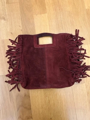 Vinröd mockaväska med fransar - Snygg handväska i vinröd mocka med rektangulärt handtag och långa fransar längs sidorna. Väskan har en mjuk, fyrkantig form och ger en boho-vibe. Perfekt accessoar för dig som vill sticka ut med detaljer och färg. Det finns ett längre band till. 