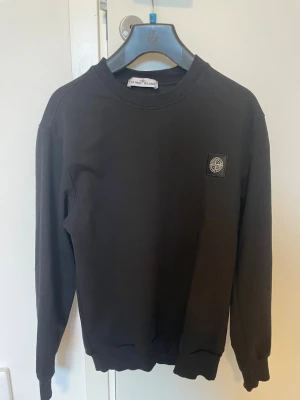 Svart sweatshirt från Stone Island - Säljer en svart sweatshirt från Stone Island med rund hals och ribbade muddar. Tröjan har en diskret logotyp-patch på bröstet och är tillverkad i mjukt bomullsmaterial. Perfekt för en clean och stilren look.