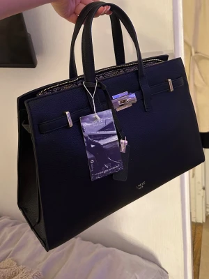 Svart handväska från LYCKE - Säljer denna superfina Lycke askim väska 🖤 Den är helt ny och aldrig använd, prislappen sitter fortfarande kvar. Väldigt stilren och rymlig modell med fina gulddetaljer. Det följer även med en längre, justerbar rem så man kan ha den över axeln eller crossover.  Köptes men blev aldrig använd, så den får gärna hitta ett nytt hem 🤍 
