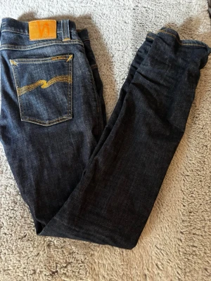 Mörkblå jeans från Nudie Jeans - Snygga mörkblå jeans från Nudie Jeans med klassisk femficksmodell och kontrastsömmar i orange. Jeansen har en rak passform och är tillverkade i kraftigt denimtyg. Bakfickan har Nudie Jeans karakteristiska broderi och läderpatch i midjan.