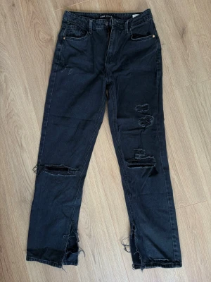 Svarta slitna jeans med hål - Säljer ett par svarta jeans med flera slitningar och hål på benen samt fransiga benslut. Jeansen har klassisk femficksmodell och rak passform. Perfekta för dig som gillar en edgy och trendig stil.