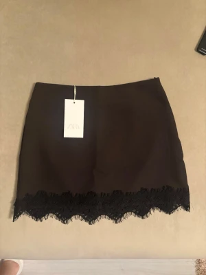 Svart minikjol med spetskant från Zara - Snygg svart minikjol från Zara med dekorativ spetskant längst ner. Kjolen har en rak passform och är tillverkad i ett mjukt tyg som ger en stilren look. Perfekt för dig som vill ha en trendig och enkel kjol med det lilla extra.