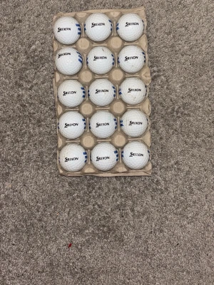 Srixon golfbollar i kartong 15-pack - Ett set med vita golfbollar från Srixon, placerade i en kartongbricka. Bollarna har klassisk dimple-yta och blå markeringar på vissa av dem. Perfekt för dig som vill fylla på golfväskan inför säsongen. 15-pack