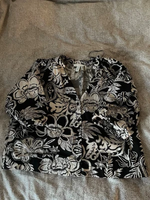Svart blommig blus med v-ringning - Snygg svart blus med stora vita blommor och v-ringning. Blusen har knappar framtill och långa ärmar med lätt puff vid axlarna. Perfekt för dig som gillar ett statement-plagg med coolt mönster. 🖤🤍