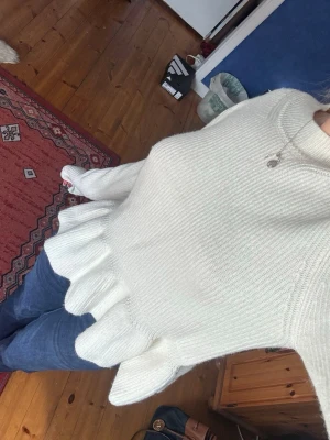 Vit stickad tröja med volang - Supersöt vit stickad tröja med volang nedtill och ribbad struktur. Tröjan har lång ärm och en rund halsringning. Perfekt för dig som vill ha en feminin och trendig look till jeans eller kjol.