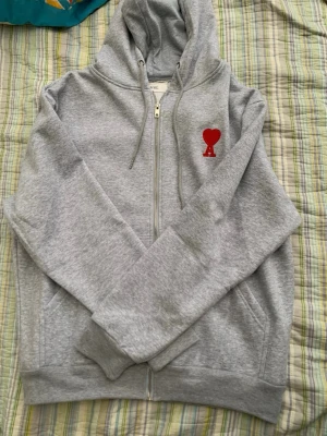 Grå hoodie med röd Ami-logo - Snygg grå zip-hoodie från Ami med huva och dragsko. På bröstet finns Ami:s ikoniska röda hjärta med bokstaven A broderat. Hoodien har fickor på sidorna och ribbade muddar. Perfekt för en chill och stilren look.