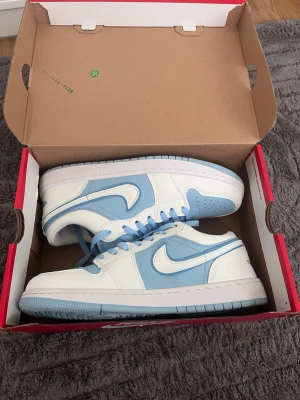 Nike Air Jordan 1 Low blå/vit - Säljer ett par Nike Air Jordan 1 Low sneakers i fräscha blå och vita färger. Skorna har klassisk snörning, vit bas med ljusblå detaljer och den ikoniska swoosh-loggan på sidan. Perfekta för dig som vill ha en clean och sportig look. Skorna passar som 43 men det står annat 