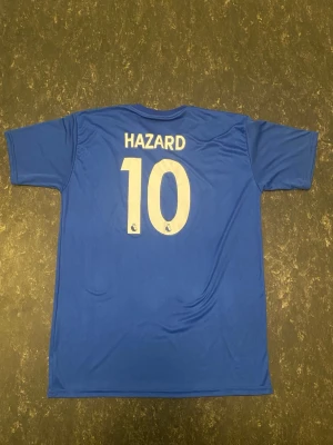 Chelsea Hazard 10 matchtröja blå - Säljer en blå Chelsea matchtröja med Hazard och nummer 10 på ryggen. Tröjan har korta ärmar, rund hals och är tillverkad i ett lätt, andningsbart material. Framtill finns Chelsea FC-logga, Nike-logga och Yokohama Tyres som sponsortryck.