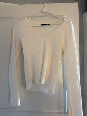 Vit ribbad långärmad topp - Säljer en stilren vit topp med ribbad struktur och v-ringning med slits längst ner på armen. Toppen har lång ärm och är tillverkad i ett mjukt, stretchigt material som sitter snyggt på kroppen.