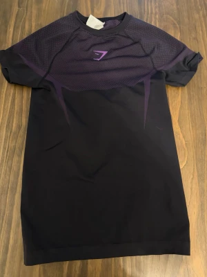 Onyx • Gymshark • lila • Small - Snygg lila och svart träningströja från Gymshark med korta ärmar och rund hals. Tröjan har ett mönstrat meshparti upptill och Gymshark-logga i lila på bröstet. Tillverkad i ett stretchigt funktionsmaterial som andas, perfekt för gymmet eller löpning.