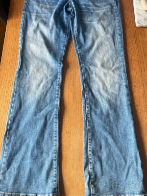 Blå bootcut jeans med slitningar - Ett par klassiska blå bootcut jeans i denim med snygga slitningar framtill. Jeansen har normal midja och utsvängda ben, vilket ger en retro och avslappnad vibe. Perfekta för dig som gillar en tidlös och trendig look.