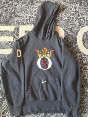 Svart Nike Hoodie med Oklahoma FC-tryck - Snygg svart hoodie med stor huva och magficka. På bröstet finns ett färgglatt Oklahoma FC-tryck med krona och bokstäverna D på ena ärmen. Nike-logga på fickan. Perfekt för dig som gillar sportig stil och vill sticka ut lite extra.
