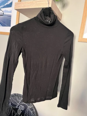 Svart polotopp med lång ärm - En svart, enkel och stilren polotopp med lång ärm. Toppen har en tight passform och är tillverkad i ett mjukt och stretchigt material, perfekt att styla till olika outfits. Polokragen ger en klassisk look och passar bra till lager-på-lager.
