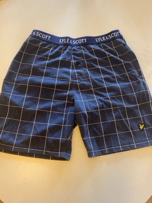 Rutiga pyjamasshorts från Lyle & Scott - Mörkblå pyjamasshorts från Lyle & Scott med vitt rutmönster och elastisk midja med logotyp. Klassisk passform, knapp framtill och mjukt material som känns skönt mot huden. Perfekta för chill hemma.