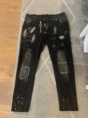 Svarta jeans med tryck och detaljer - Unika svarta jeans med coola vita och gula färgstänk, slitningar och trycket 'BE COOL BE NICE' baktill. Dragkedja framtill med ringdetalj. Raka ben och femficksmodell. Perfekta för dig som vill sticka ut med din stil.
