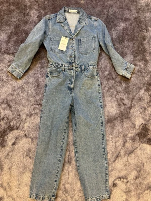 Blå denim jumpsuit från ZARA - Snygg blå denim jumpsuit från ZARA med klassisk skjortkrage, långa ärmar och raka ben. Två bröstfickor och fickor vid höfterna. Stängs med knappar framtill och har bälteshällor i midjan. Perfekt för en cool och avslappnad look.