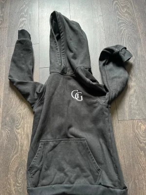 Svart hoodie med GG-logga - Säljer en svart hoodie med stor huva och en vit broderad GG-logga på bröstet. Hoodien har en klassisk känguruficka framtill och ribbade muddar vid ärmslut och nederkant. Perfekt för en chill och avslappnad stil.