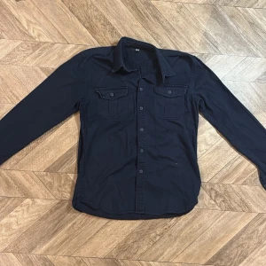 Mörkblå UNIQLO skjorta - Stilren mörkblå skjorta med långa ärmar och två bröstfickor med lock och knapp. Klassisk krage och knäppning framtill. Skjortan har en enkel och tidlös design som passar till många olika stilar. Materialet ser ut att vara bomull eller en bomullsblandning.