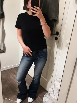 Blå bootcut jeans med slitningar - Säljer ett par blå bootcut jeans med slitningar på knäna och rå kant nedtill. Jeansen har en klassisk femficksmodell och tydliga sömmar på bakfickorna. Perfekta för en avslappnad och trendig look.