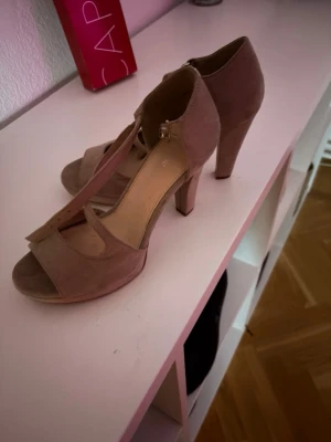 Beige/rosa  peep toe pumps med T-rem - Säljer ett par snygga beige pumps med hög klack och öppen tå. Skorna har en klassisk T-rem. Materialet ser ut att vara mockaimitation och de har en stilren design som passar till många outfits. Säljer dessa då jag inte använder dem, praktiskt sett i nyskick.