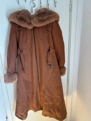 Brun kappa med fuskpäls och huva - Lång brun kappa med stor huva och mjuka fuskpälsdetaljer vid krage och ärmslut. Kappan har dragkedja framtill, dekorativa band i midjan och bälte bak. Materialet ser ut som ullmix och modellen är A-linjeformad för en snygg siluett.