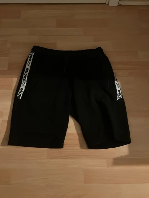 Svarta shorts med vita detaljer - Säljer ett par svarta shorts med vita band längs sidorna där det står 'LOWD'. Shortsen har snörning i midjan och dragkedjeförsedda fickor. Perfekta för en avslappnad stil och funkar till både chill och aktivitet.