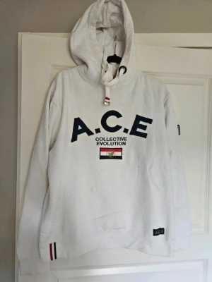 Vit hoodie med A.C.E tryck - Vit hoodie från A.C.E Collective Evolution XL  Vit hoodie från A.C.E Collective Evolution med broderad logga och text i mörkblått på bröstet. Tröjan har huva med snörning, ribbade muddar och detaljer i rött, vitt och blått. Snygg patch på ärmen och nertill. Perfekt för dig som gillar streetwear.  Osäker på om det är herr eller dam storlek. Tror herr !!!!Liten i storlek!!!!!!  Kan även hämtas I Partille  E1