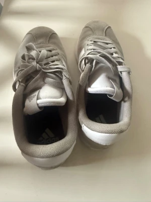 Addias -VL Court  sneakers  - Säljer ett par vita Adidas sneakers med grå detaljer och klassiska ränder på sidan. Skorna har låga skaft och är tillverkade i mocka och textil. Perfekta för dig som gillar en clean och sportig look.