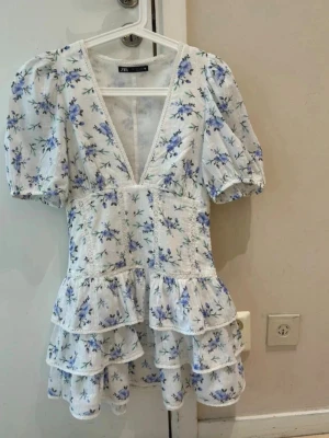  Zara Vit blommig kortklänning med volanger - Superfin vit kortklänning med blått blommönster, djup v-ringning och puffärmar. Klänningen har flera volanglager och är perfekt för sommarens alla tillfällen. Tillverkad i ett lätt och luftigt material som känns skönt mot huden.