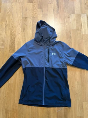 Under Armour blå och grå vindjacka - Snygg vindjacka från Under Armour i tvåfärgad design med mörkblå nederdel och ärmar samt grå överdel och huva. Jackan har dragkedja framtill, bröstficka med dragkedja och en vit logga på bröstet. Perfekt för blåsiga dagar.