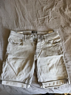Grå jeansshorts från Jack & Jones - Snygga grå jeansshorts från Jack & Jones med klassisk femficksdesign och uppvikta benslut. Regular fit och stängs med knappar framtill. Perfekta för varma dagar och passar till det mesta i garderoben.