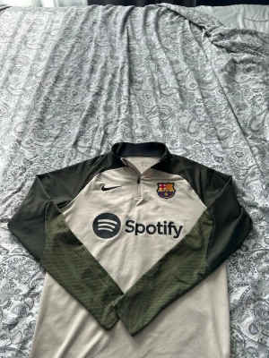 Nike Barcelona träningströja beige/grön - Snygg långärmad träningströja från Nike med FC Barcelona-logga och Spotify sponsortryck. Tröjan är beige med mörkgröna ärmar och har halv dragkedja framtill. Perfekt för fotbollsträning eller som sportig vardagsstil.