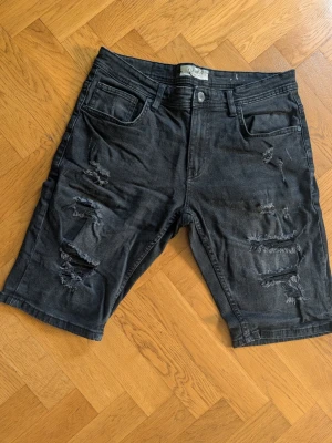 Mörka slitna jeansshorts - Säljer ett par mörka jeansshorts med slitna detaljer och flera revor framtill. Klassisk femficksmodell och midwaist passform. Materialet är bomull och de har en loose fit som ger en avslappnad vibe. Perfekta för sommaren!