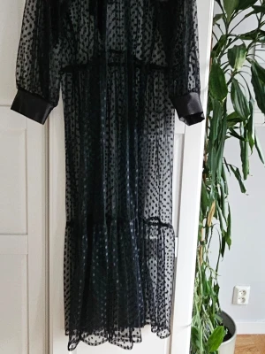 Mesh långklänning med knyt - Säljer en svart långklänning i transparent mesh med prickigt mönster. Klänningen har långa ärmar med breda manschetter och en snygg knytning vid halsen. Perfekt för dig som vill ha en statement piece med coola detaljer. Ny