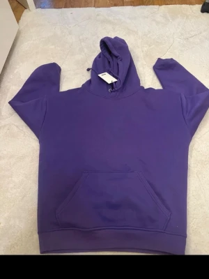 Lila hoodie med huva och magficka - Säljer en stilren lila hoodie med huva. Hoodien har en stor magficka framtill och ribbade muddar vid ärmslut och nederkant. Perfekt för dig som gillar en enkel men snygg look. Materialet känns mjukt och bekvämt. Aldrig använd i storlek xs