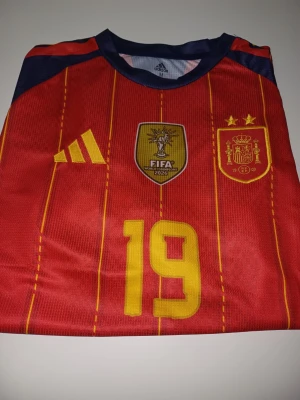 Lamine Yamal WC tröja - Röd matchtröja från Adidas för Spaniens landslag med gula detaljer, nummer 19 på framsidan och officiella FIFA-märket. Tröjan har tunna gula ränder, två stjärnor ovanför klubbmärket och mörkblå detaljer vid axlarna. Perfekt för fotbollsfans!
