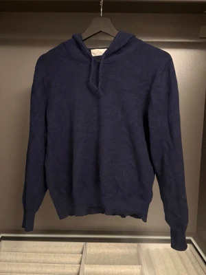 Mörkblå hoodie i kashmir - Mysig mörkblå hoodie i mjukt kashmirmaterial med huva och långa ärmar. Enkel och stilren design utan tryck eller detaljer, perfekt för en avslappnad look. Dragsko i huvan för justerbar passform. Väldig fin Kashmir tröja men lite trasig vid fram krage som man inte märker av och som lätt går att fixa bättre 
